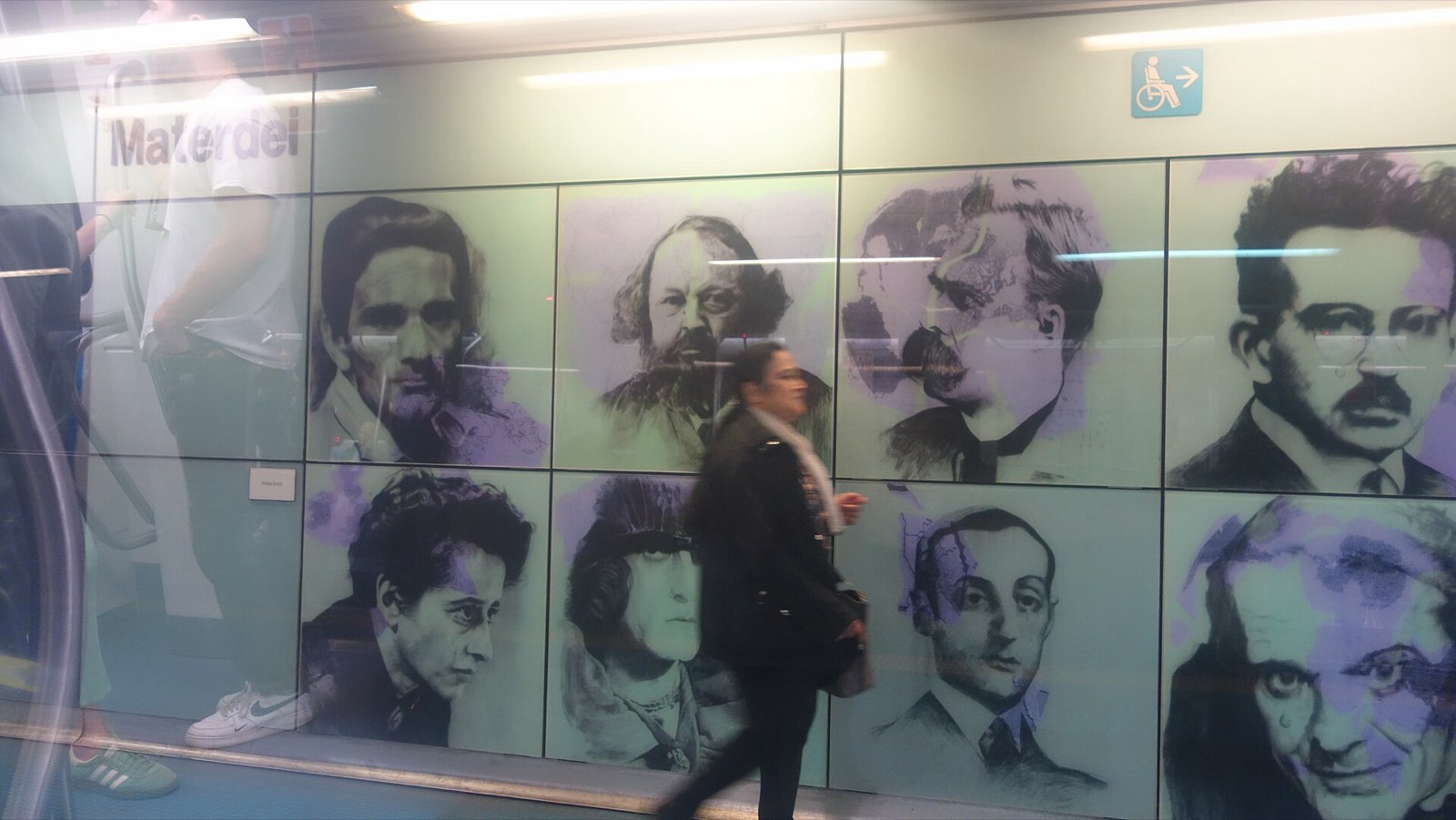 A wall of large printed portraits at Materdei station with a woman walking past